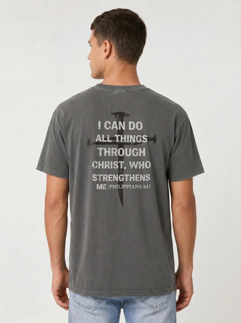 A man viewed from the back wearing a dark gray t-shirt with a white printed bible verse and cross graphic on the back, paired with light blue jeans, against a light gray studio background.