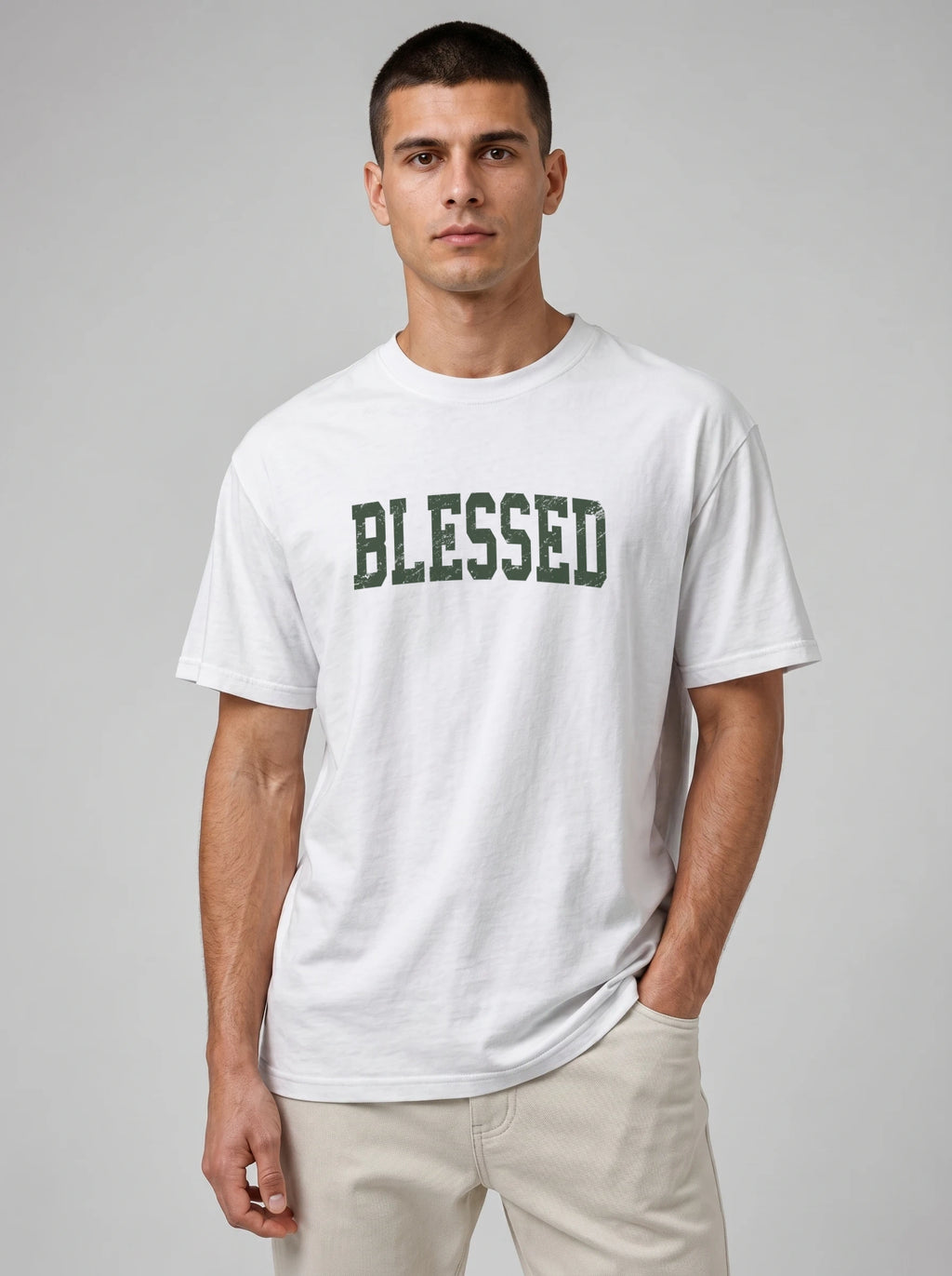 A man wearing a white crewneck t-shirt with 'BLESSED' printed in dark green varsity-style letters across the chest, paired with light beige pants, against a solid gray studio background.