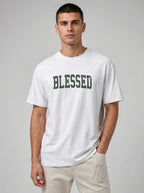 A man wearing a white crewneck t-shirt with 'BLESSED' printed in dark green varsity-style letters across the chest, paired with light beige pants, against a solid gray studio background.