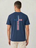 A man viewed from the back wearing a navy blue t-shirt with a distressed American flag cross graphic on the back, paired with beige pants, against a light gray studio background.