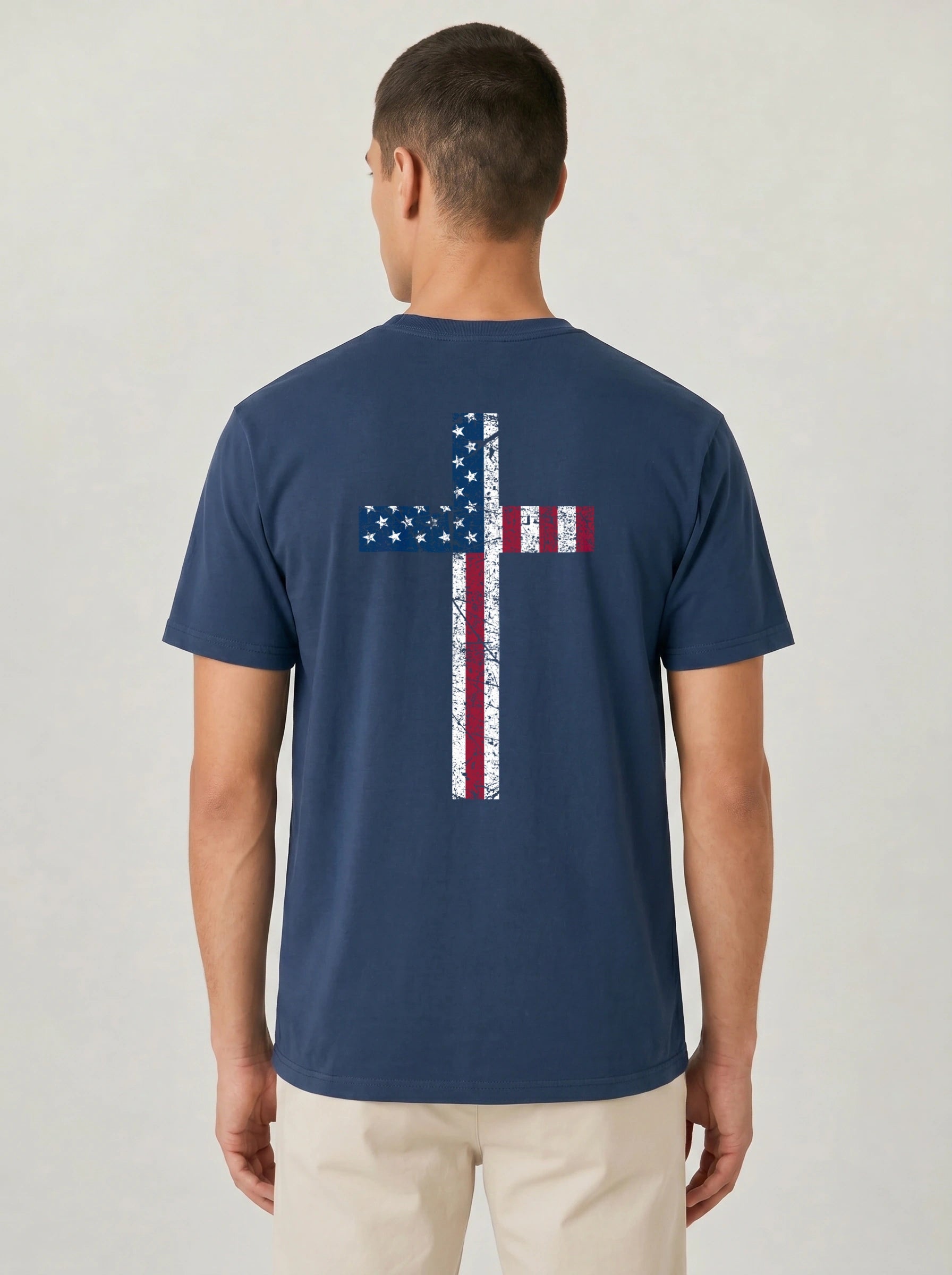 A man viewed from the back wearing a navy blue t-shirt with a distressed American flag cross graphic on the back, paired with beige pants, against a light gray studio background.