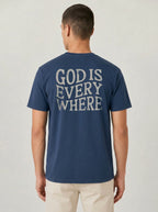 A man viewed from the back wearing a navy blue t-shirt with 'GOD IS EVERY WHERE' printed in a distressed white font across the back, paired with light beige pants, against a light gray studio background.