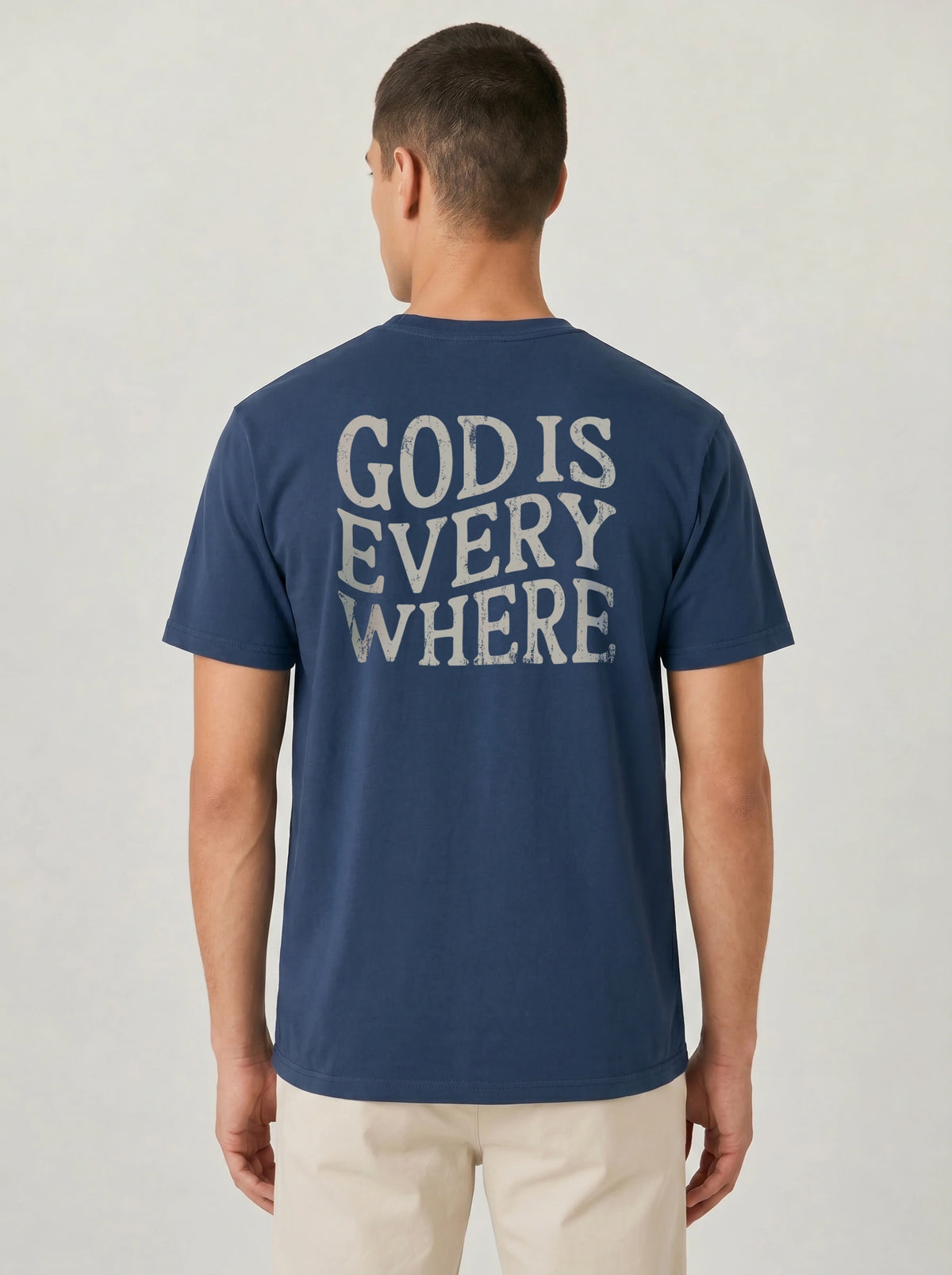 A man viewed from the back wearing a navy blue t-shirt with 'GOD IS EVERY WHERE' printed in a distressed white font across the back, paired with light beige pants, against a light gray studio background.