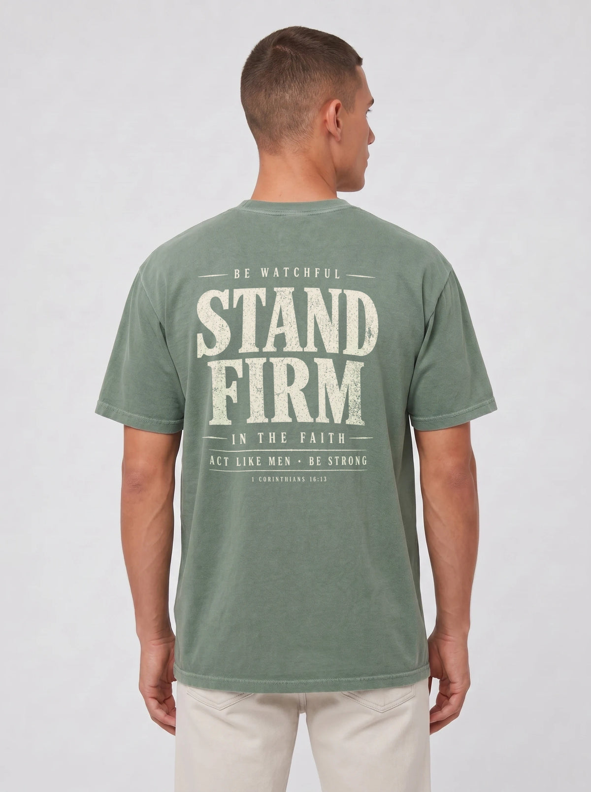 A man viewed from the back wearing a sage green t-shirt with a distressed white graphic print that reads 'STAND FIRM' and 'BE WATCHFUL IN THE FAITH'.