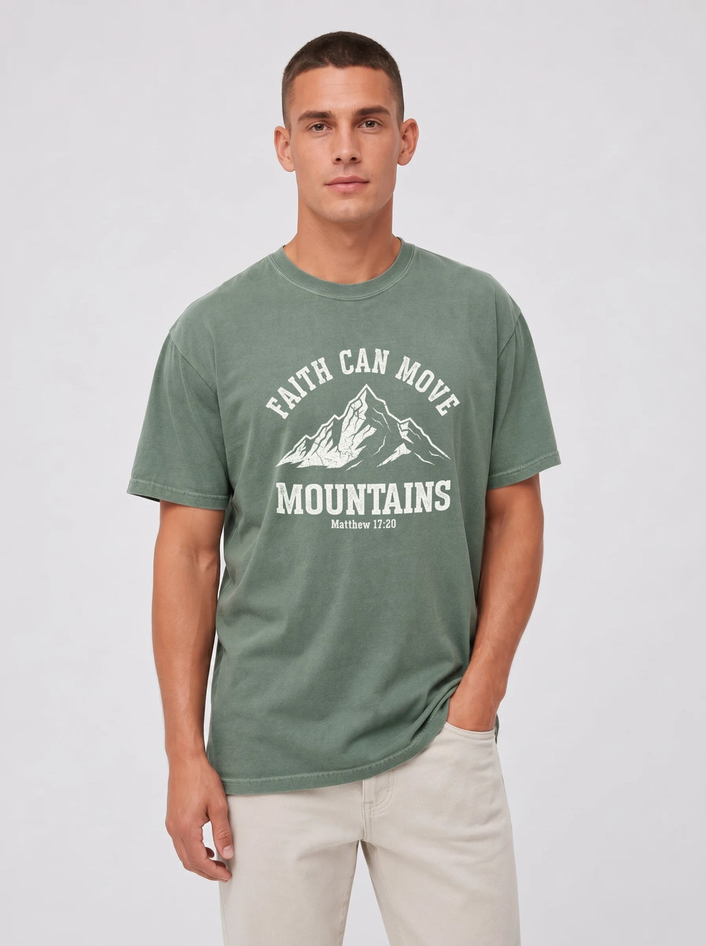 A man wearing a sage green graphic t-shirt with a white mountain print and the text 'FAITH CAN MOVE MOUNTAINS' on the front, paired with light beige pants, against a light gray studio background.