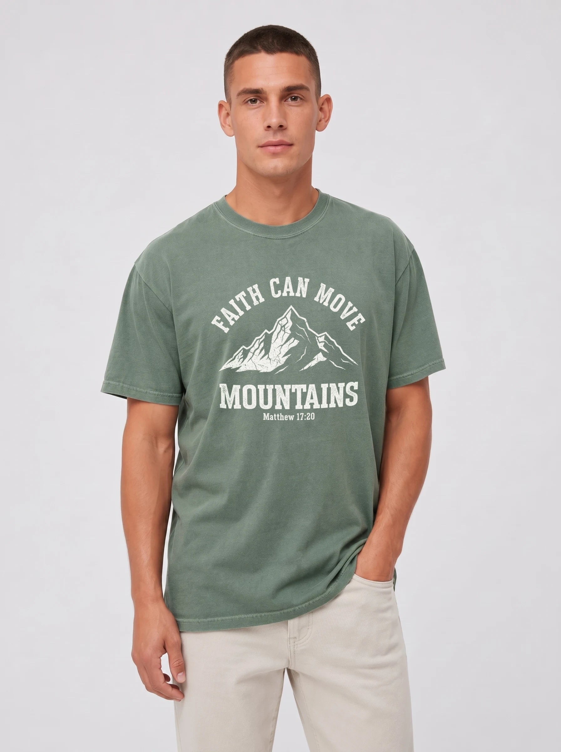 A man wearing a sage green graphic t-shirt with a white mountain print and the text 'FAITH CAN MOVE MOUNTAINS' on the front, paired with light beige pants, against a light gray studio background.