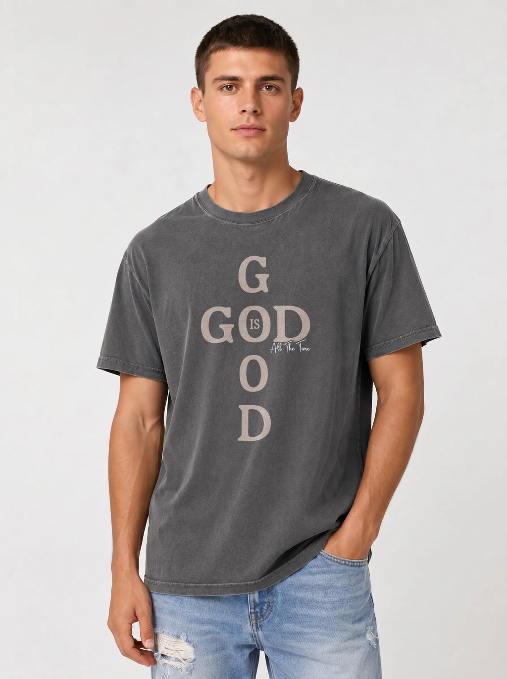 A man wearing a dark gray washed graphic t-shirt with 'GOD' printed vertically in a distressed beige font, paired with light blue jeans, against a light gray studio background.