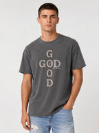 A man wearing a dark gray washed graphic t-shirt with 'GOD' printed vertically in a distressed beige font, paired with light blue jeans, against a light gray studio background.