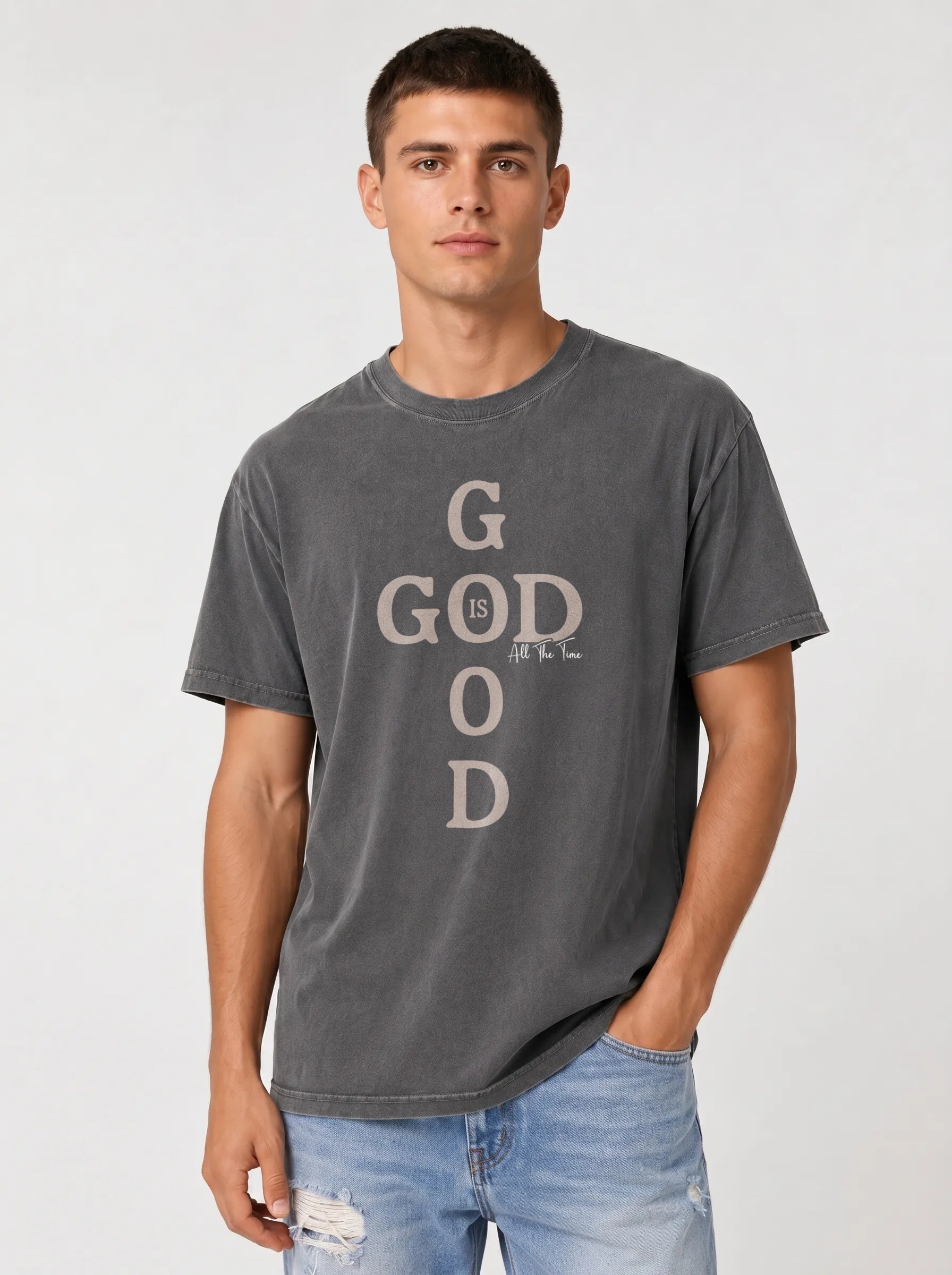 A man wearing a dark gray washed graphic t-shirt with 'GOD' printed vertically in a distressed beige font, paired with light blue jeans, against a light gray studio background.