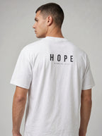 A man wearing a white short-sleeve t-shirt, viewed from the back. The shirt features the word 'HOPE' printed in black capital letters across the upper back, with 'ROMANS 15:13' printed underneath in smaller text.