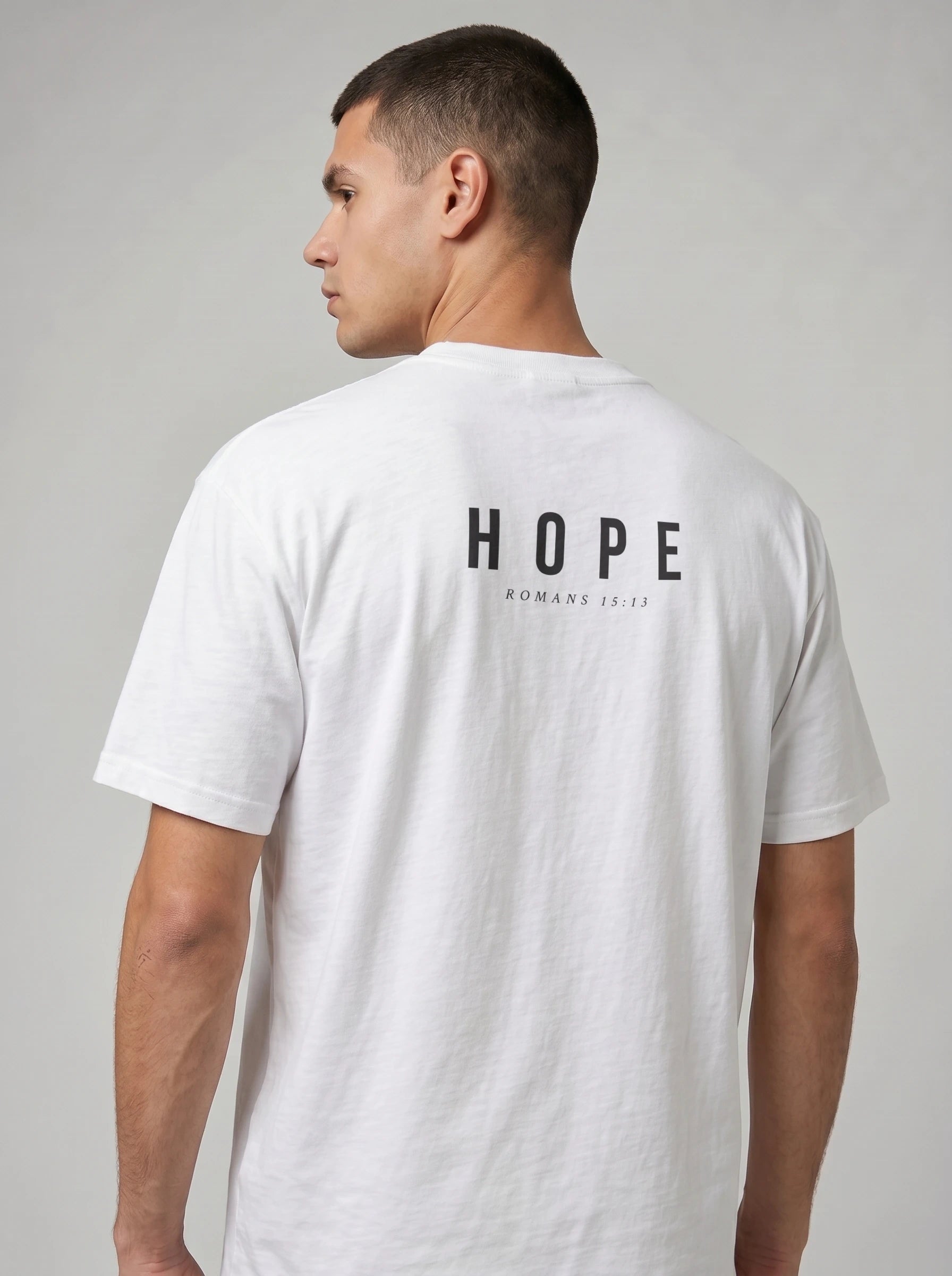 A man wearing a white short-sleeve t-shirt, viewed from the back. The shirt features the word 'HOPE' printed in black capital letters across the upper back, with 'ROMANS 15:13' printed underneath in smaller text.