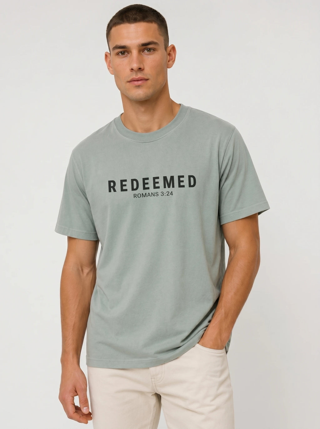 A man wearing a sage green graphic t-shirt with 'REDEEMED ROMANS 3:24' printed in black across the chest, paired with off-white trousers, against a light gray studio background.