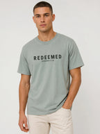 A man wearing a sage green graphic t-shirt with 'REDEEMED ROMANS 3:24' printed in black across the chest, paired with off-white trousers, against a light gray studio background.