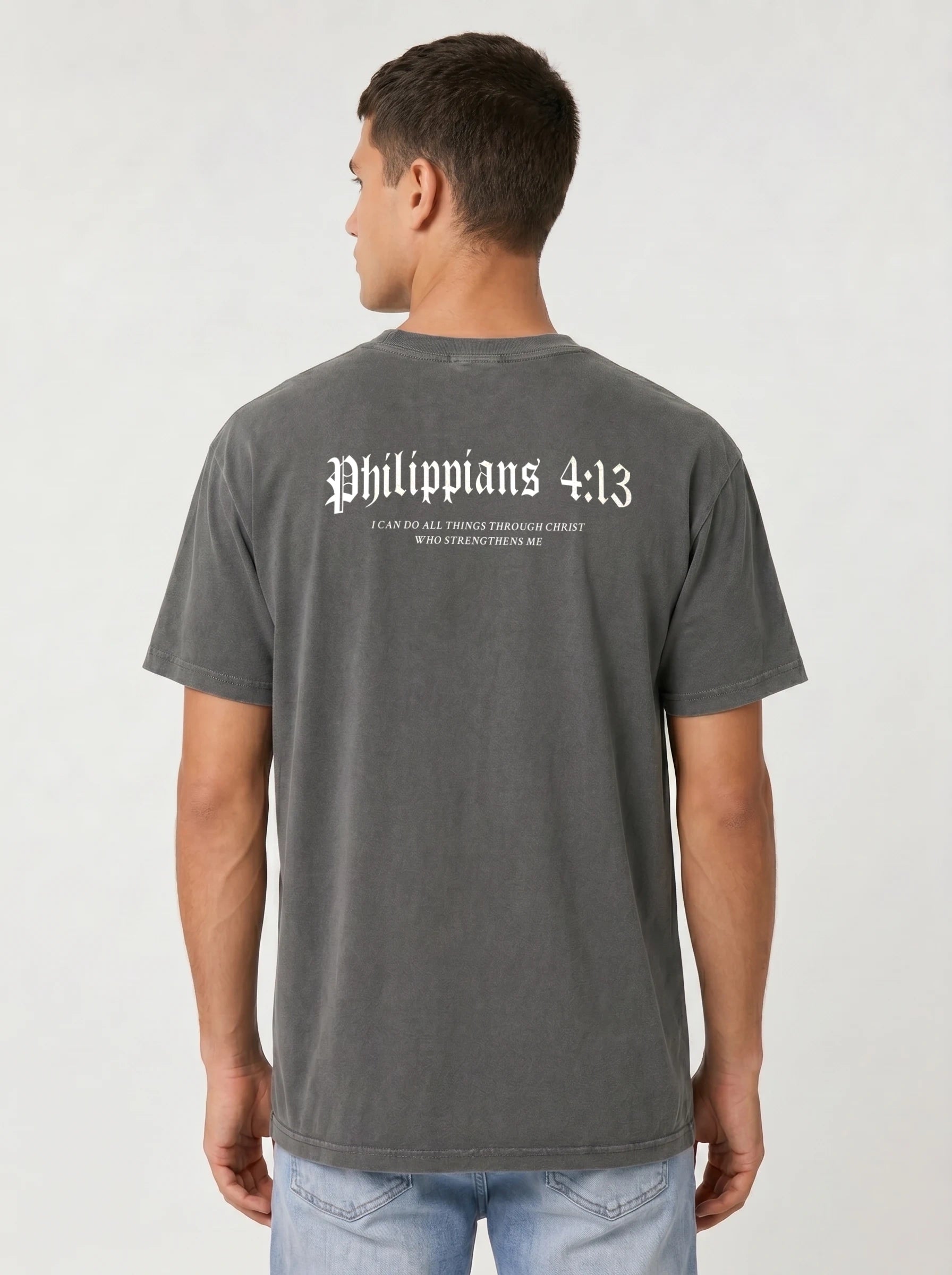 A man wearing a dark gray washed t-shirt from the back, featuring the text 'Philippians 4:13' in white gothic font with smaller scripture text underneath. He is also wearing light blue jeans, photographed against a neutral light gray background.