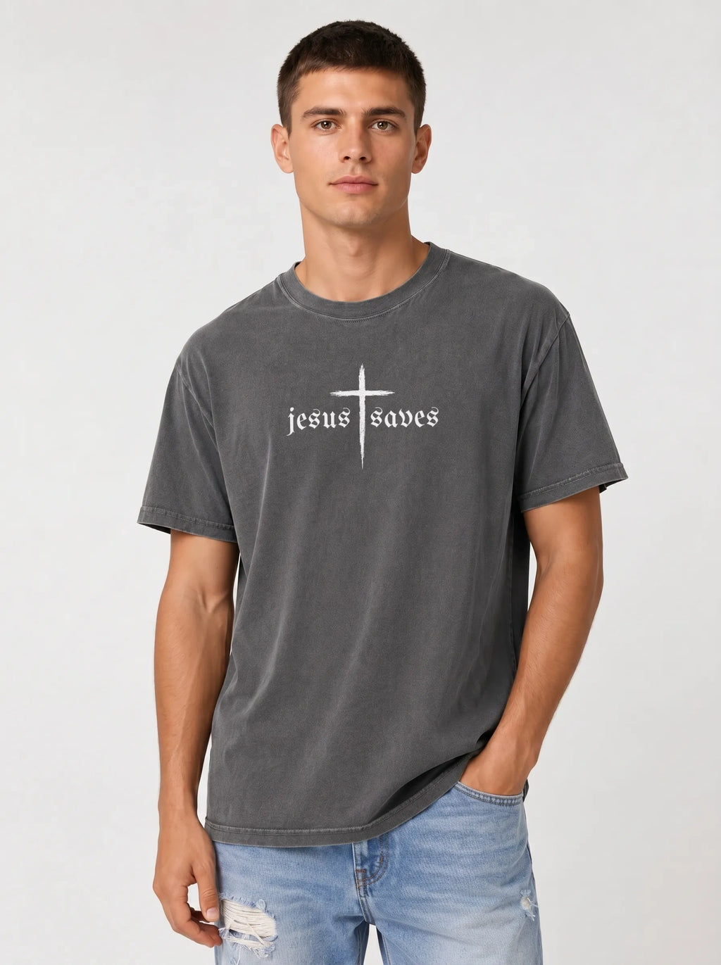 A man wearing a dark gray washed t-shirt with a white graphic that reads 'jesus saves' under a cross, paired with light blue jeans, against a light gray studio background.