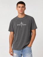 A man wearing a dark gray washed t-shirt with a white graphic that reads 'jesus saves' under a cross, paired with light blue jeans, against a light gray studio background.