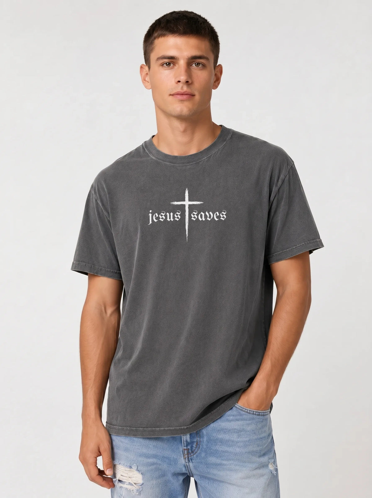 A man wearing a dark gray washed t-shirt with a white graphic that reads 'jesus saves' under a cross, paired with light blue jeans, against a light gray studio background.