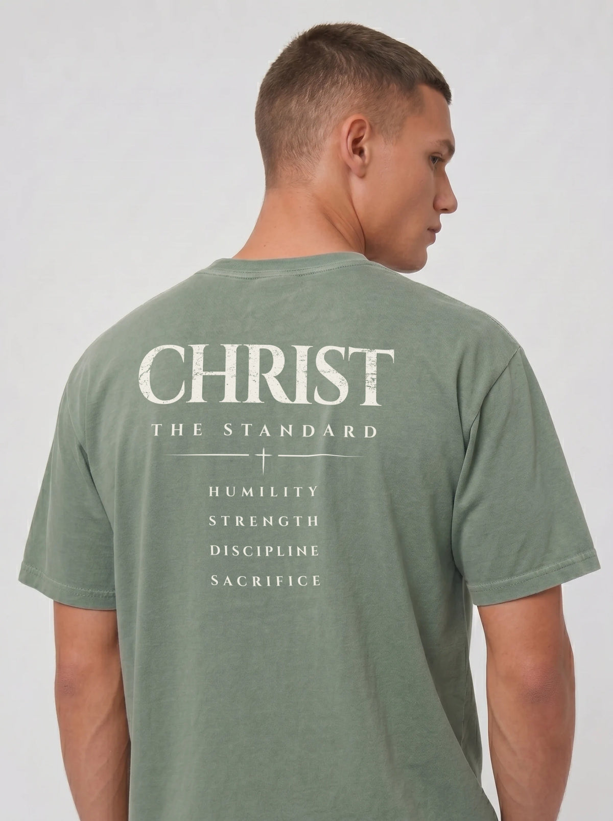 Person wearing a muted green T-shirt with “Christ The Standard,” a small cross, and “Humility, Strength, Discipline, Sacrifice” on the back.
