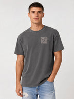 A man wearing a dark gray washed t-shirt with the text 'GOD IS EVERY WHERE' printed in white on the left chest, paired with light blue jeans, against a light gray studio background.
