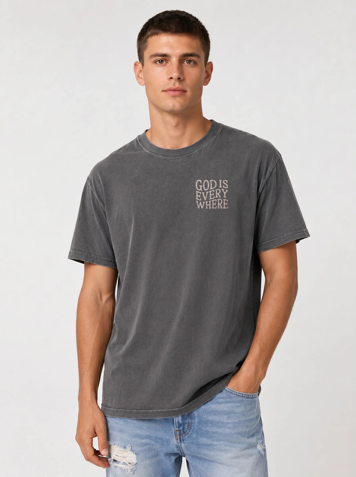 A man wearing a dark gray washed t-shirt with the text 'GOD IS EVERY WHERE' printed in white on the left chest, paired with light blue jeans, against a light gray studio background.