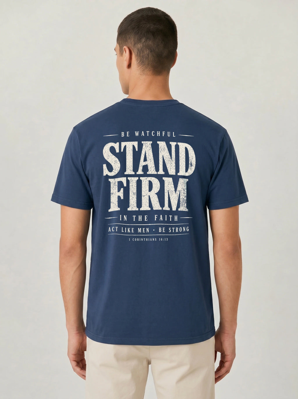 A man viewed from behind wearing a navy blue t-shirt with a large distressed white graphic on the back that reads 'STAND FIRM' with smaller text above and below, paired with light beige pants against a light gray studio background.