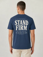 A man viewed from behind wearing a navy blue t-shirt with a large distressed white graphic on the back that reads 'STAND FIRM' with smaller text above and below, paired with light beige pants against a light gray studio background.