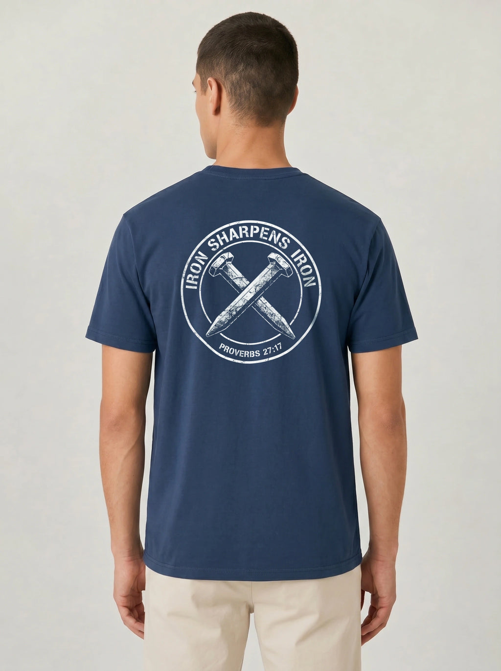 A man viewed from the back wearing a navy blue t-shirt with a large white circular graphic on the back that reads 'IRON SHARPENS IRON' and 'PROVERBS 27:17' around crossed hammers. He is also wearing light beige pants, set against a plain light gray studio background.