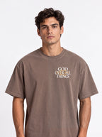 Person wearing a washed brown T-shirt with a small chest graphic reading “God Over All Things.”
