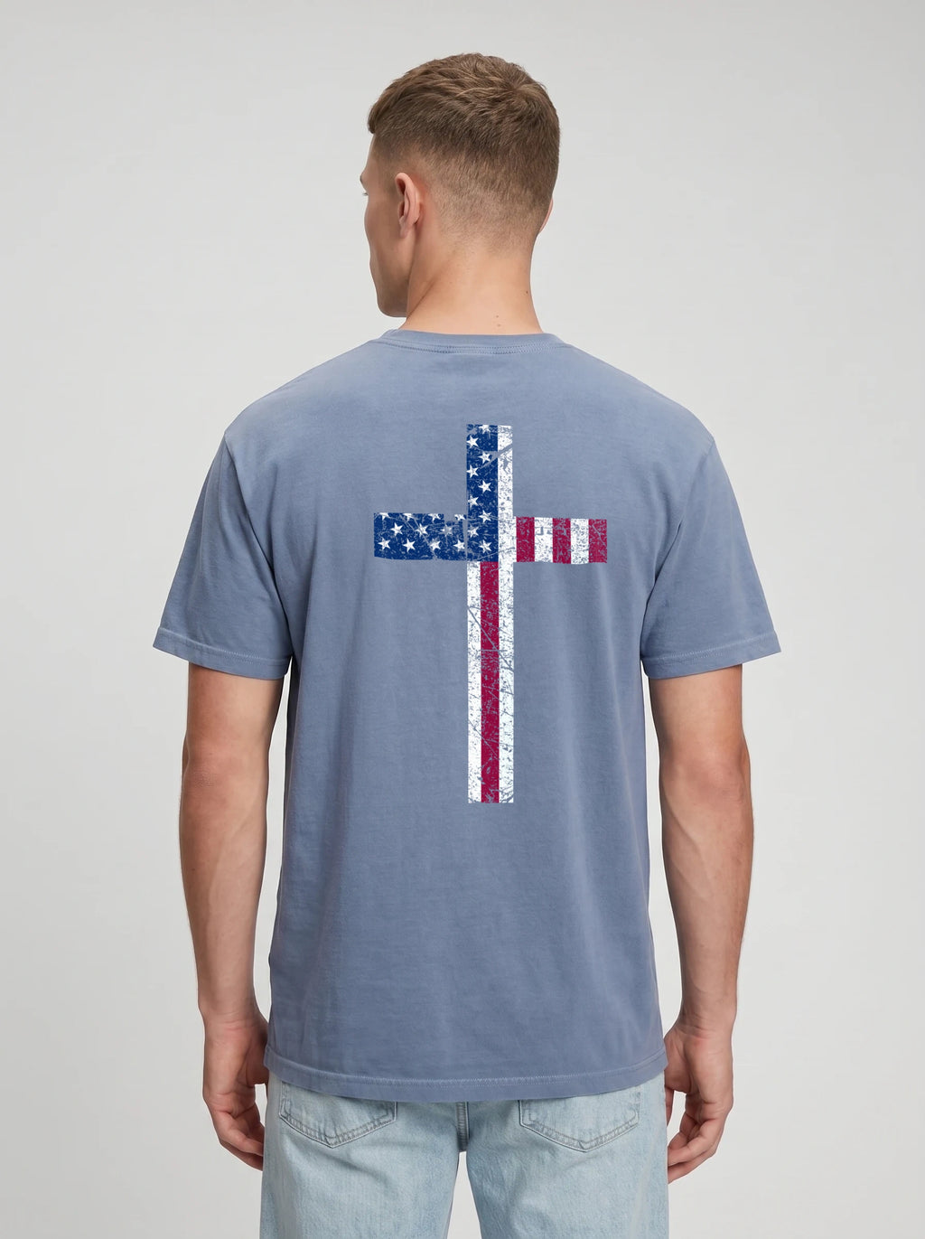 A man viewed from the back wearing a slate blue t-shirt with a distressed American flag cross graphic on the back, paired with light wash jeans, against a light gray studio background.
