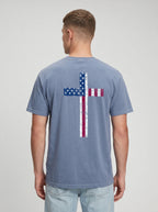 A man viewed from the back wearing a slate blue t-shirt with a distressed American flag cross graphic on the back, paired with light wash jeans, against a light gray studio background.