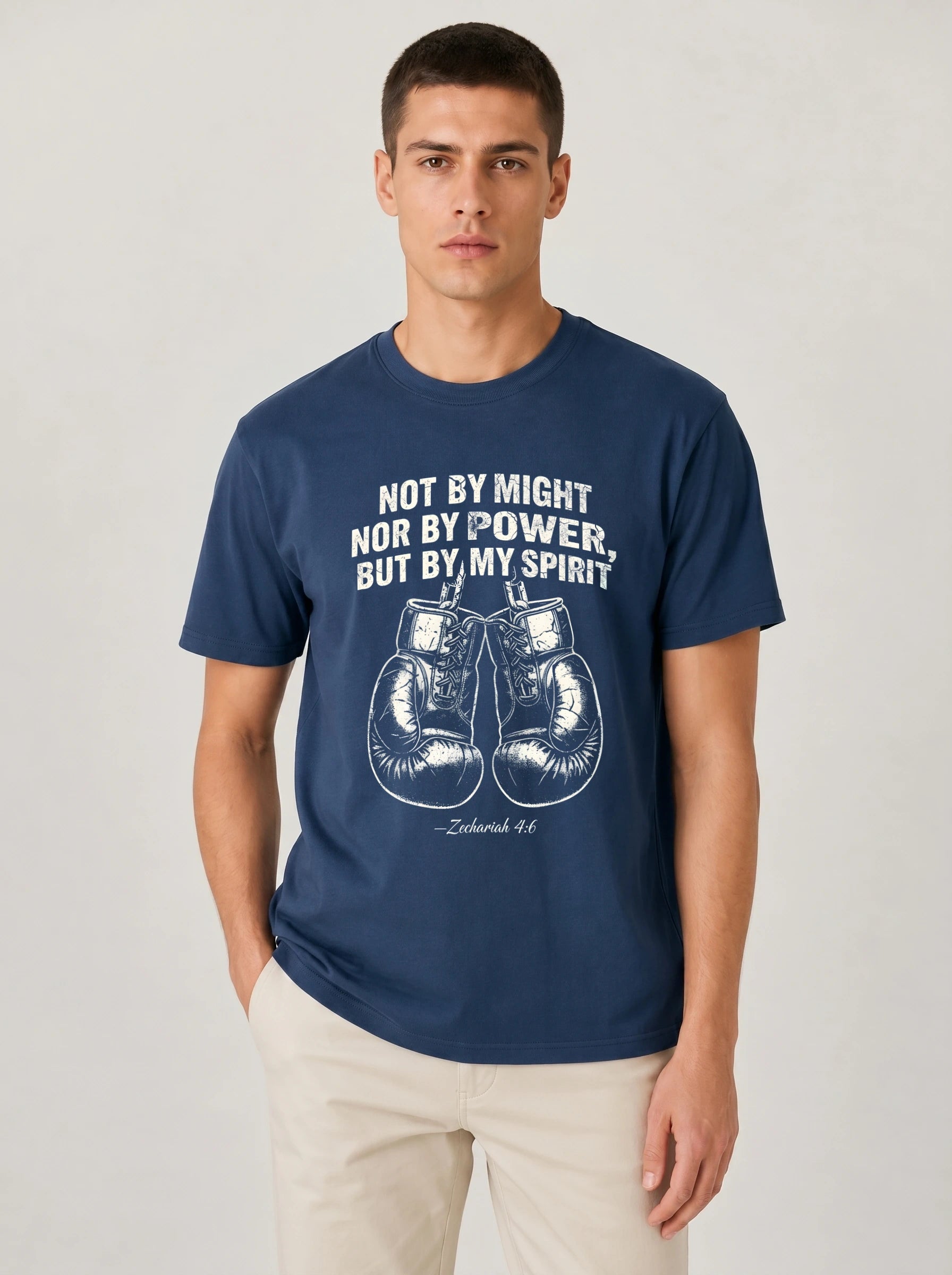 A man wearing a navy blue t-shirt with a white graphic print of boxing gloves and the quote 'NOT BY MIGHT NOR BY POWER, BUT BY MY SPIRIT' on a light gray studio background.