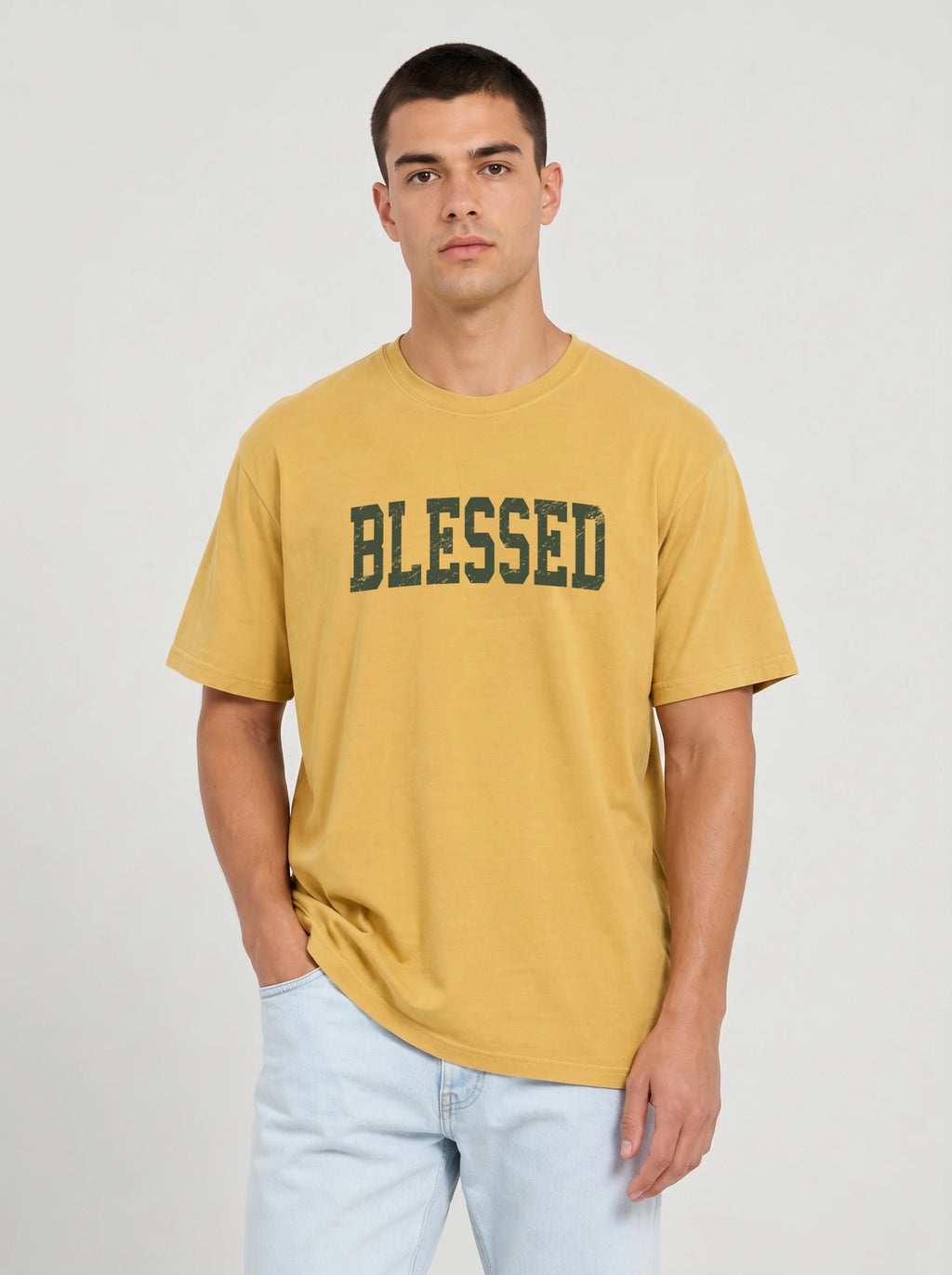 A man wearing a mustard yellow t-shirt with 'BLESSED' printed in dark green collegiate-style letters across the chest, paired with light blue jeans, against a light gray studio background.
