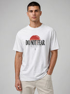 A man wearing a white graphic t-shirt with a red sun and black text reading 'DO NOT FEAR' and 'I AM WITH YOU', paired with beige pants, against a solid gray studio background.