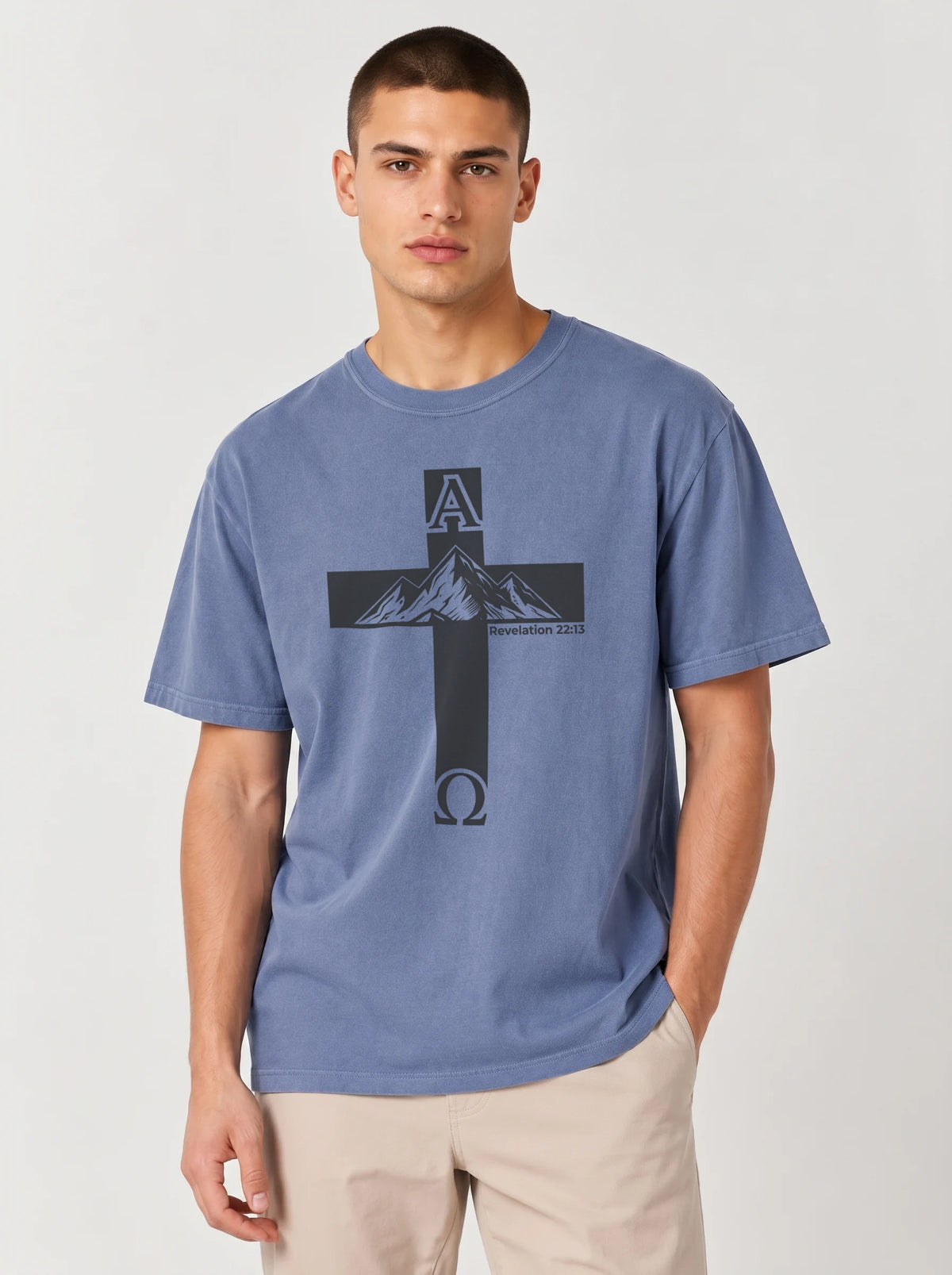 A man wearing a heathered blue crewneck t-shirt with a black cross graphic featuring mountain peaks and the letters A and Omega, paired with beige pants, against a light gray studio background.