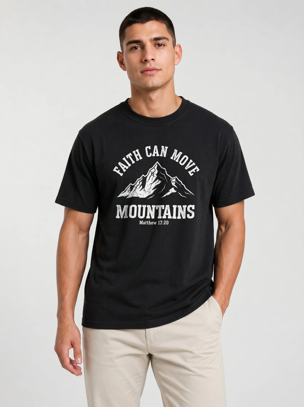A man wearing a black short-sleeve graphic t-shirt with a white mountain print and the text 'FAITH CAN MOVE MOUNTAINS' and 'Matthew 17:20', paired with beige pants, against a light gray studio background.