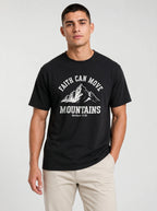 A man wearing a black short-sleeve graphic t-shirt with a white mountain print and the text 'FAITH CAN MOVE MOUNTAINS' and 'Matthew 17:20', paired with beige pants, against a light gray studio background.