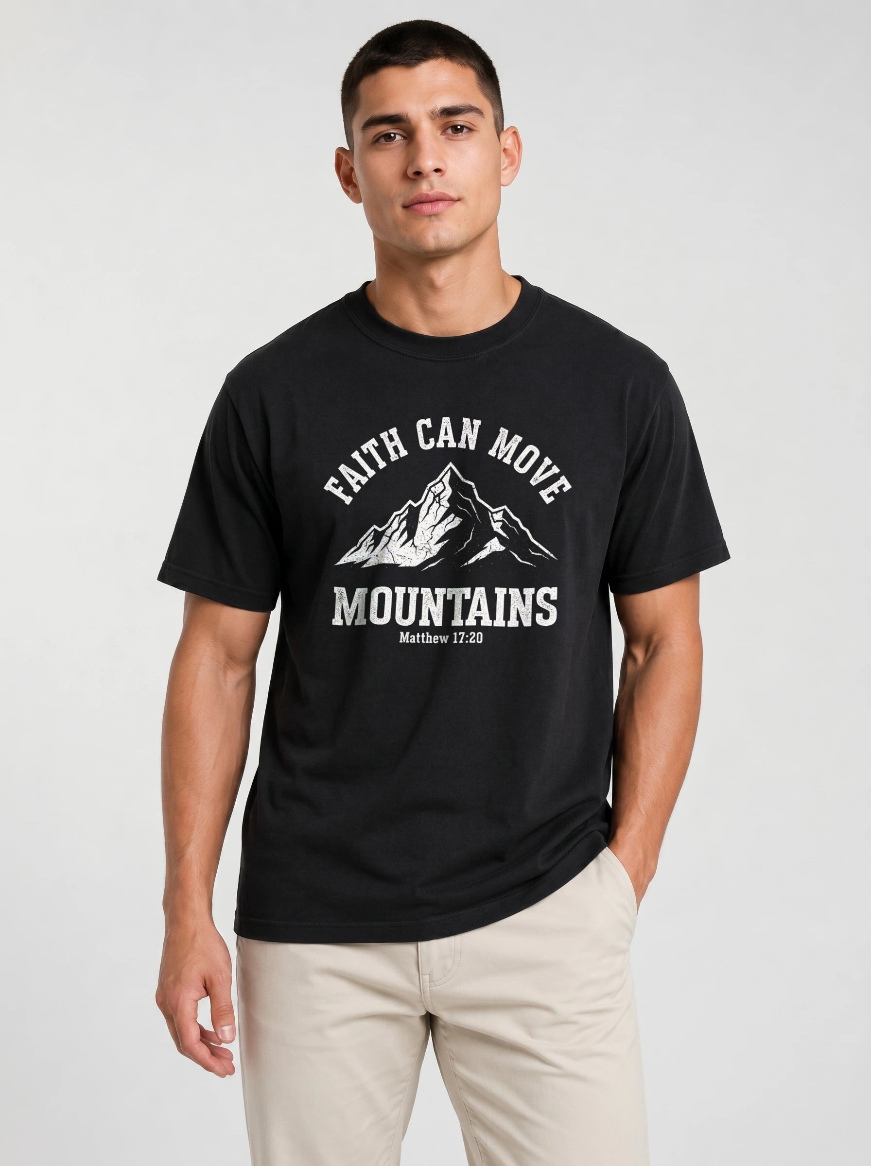 A man wearing a black short-sleeve graphic t-shirt with a white mountain print and the text 'FAITH CAN MOVE MOUNTAINS' and 'Matthew 17:20', paired with beige pants, against a light gray studio background.