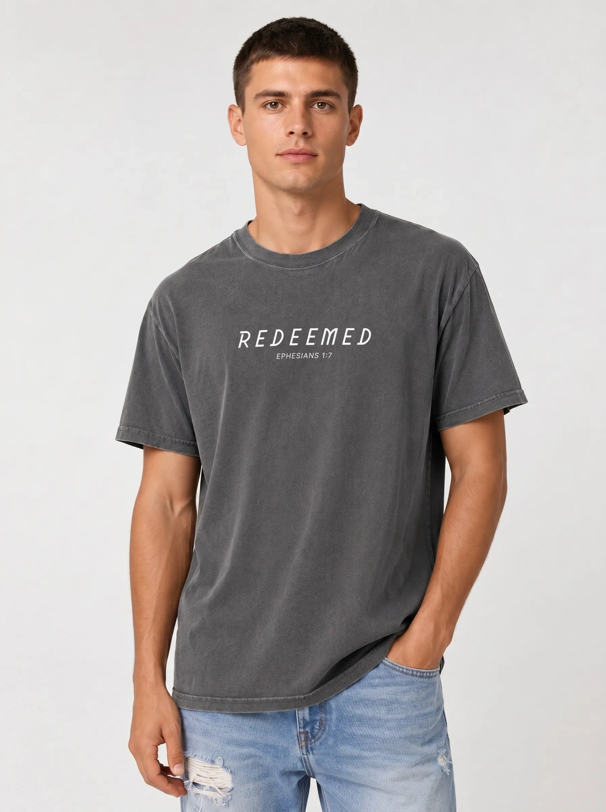 A man wearing a heather gray Comfort Colors t-shirt with "REDEEMED" in bold white capital letters and "EPHESIANS 1:7" underneath, paired with distressed blue jeans, against a plain light gray studio background.