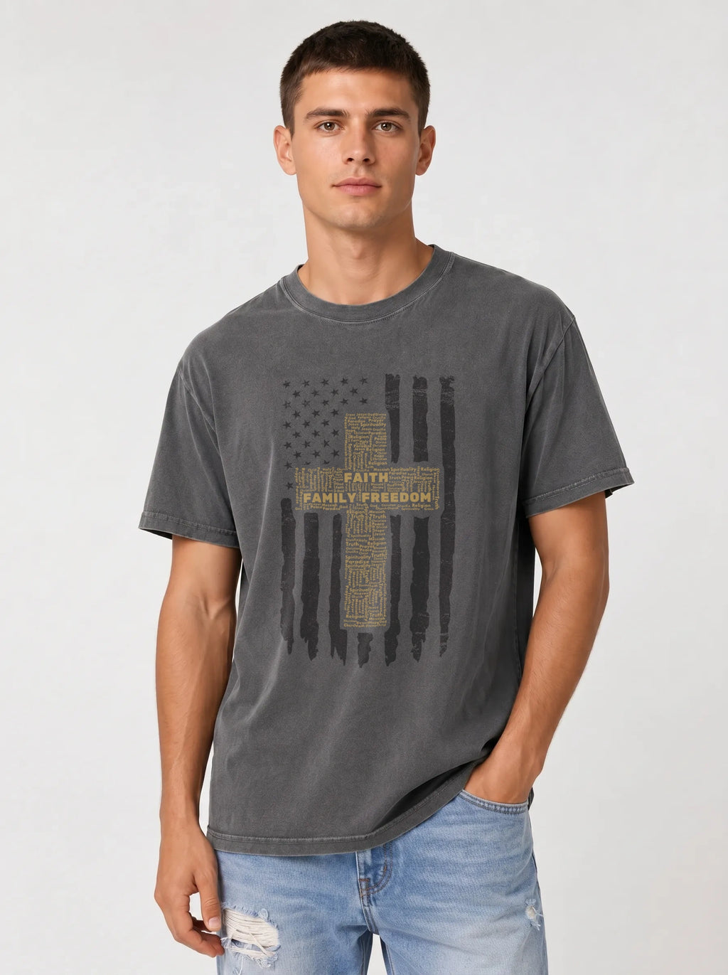 A man wearing a dark gray washed graphic t-shirt with a distressed American flag and cross design featuring the words 'FAITH FAMILY FREEDOM', paired with light blue jeans, against a light gray studio background.