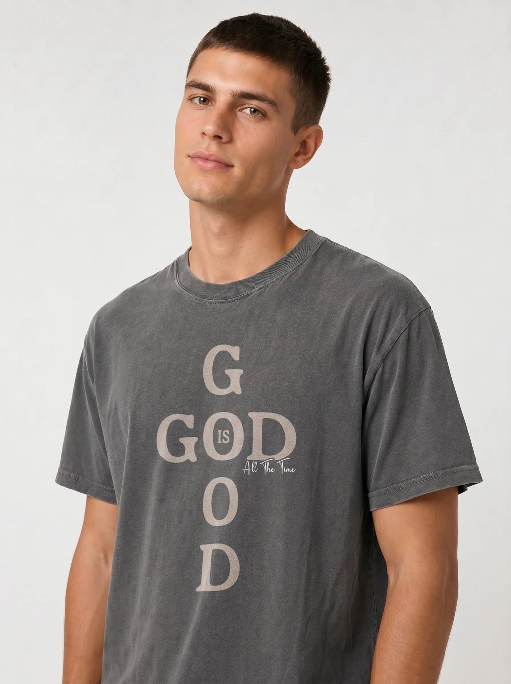 A man wearing a dark gray washed graphic t-shirt with the text 'GOD IS ALL YOU NEED' printed vertically in a light beige color on the front, set against a plain light gray background.