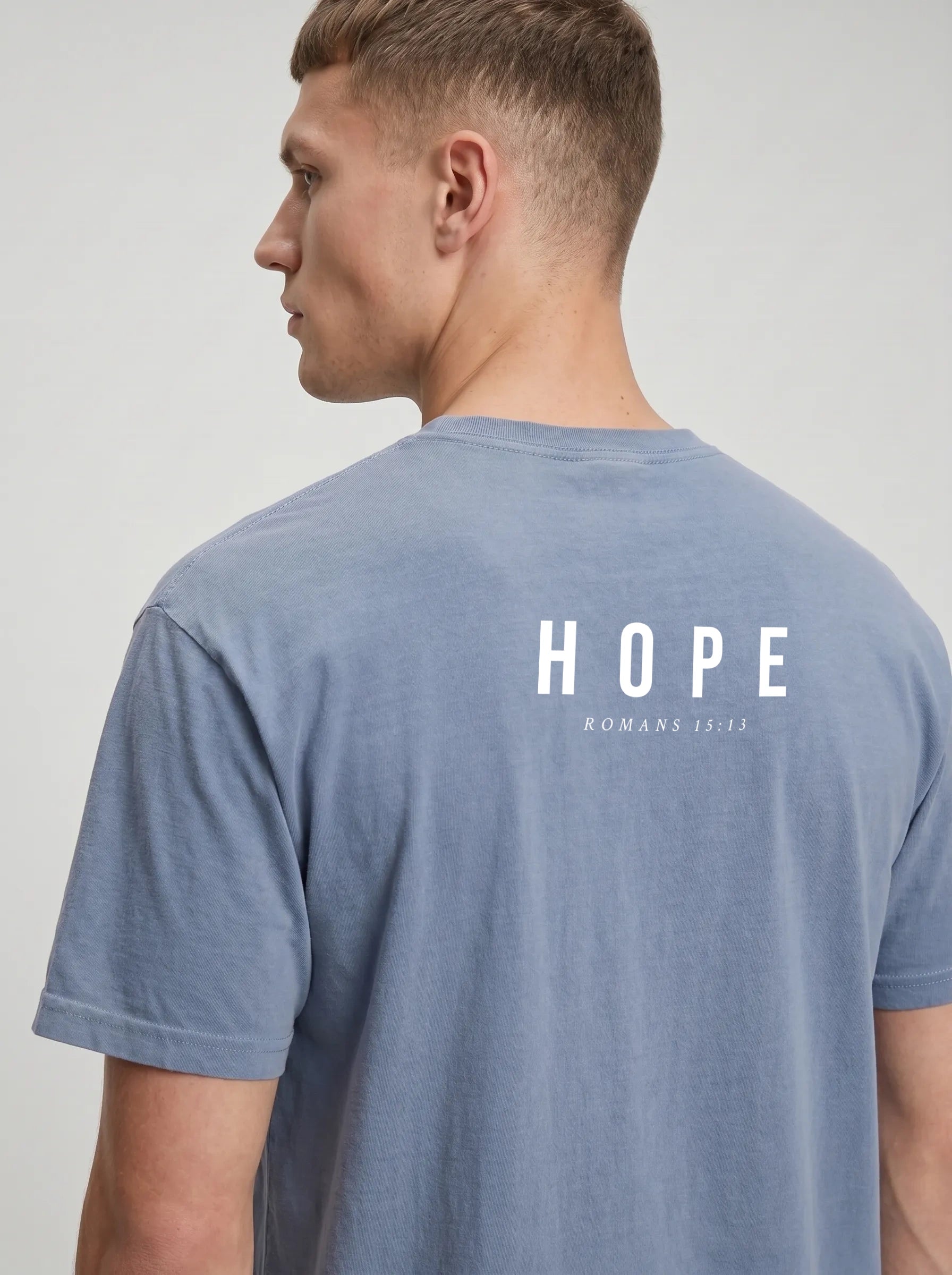 A man shown from the back wearing a heathered blue-gray t-shirt with 'HOPE ROMANS 15:13' printed in white on the back, against a light gray studio background.