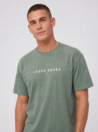 A man wearing a muted sage green t-shirt with the text 'JESUS SAVES' and a small cross printed in white on the chest, set against a plain light gray background.