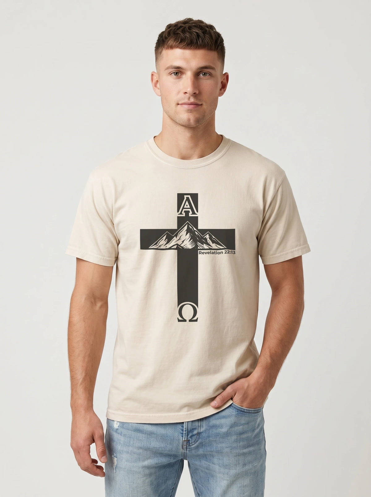 A man wearing a light beige crewneck t-shirt with a black graphic of a cross containing mountain peaks and the Greek letters Alpha and Omega, paired with light blue jeans, against a light gray background.