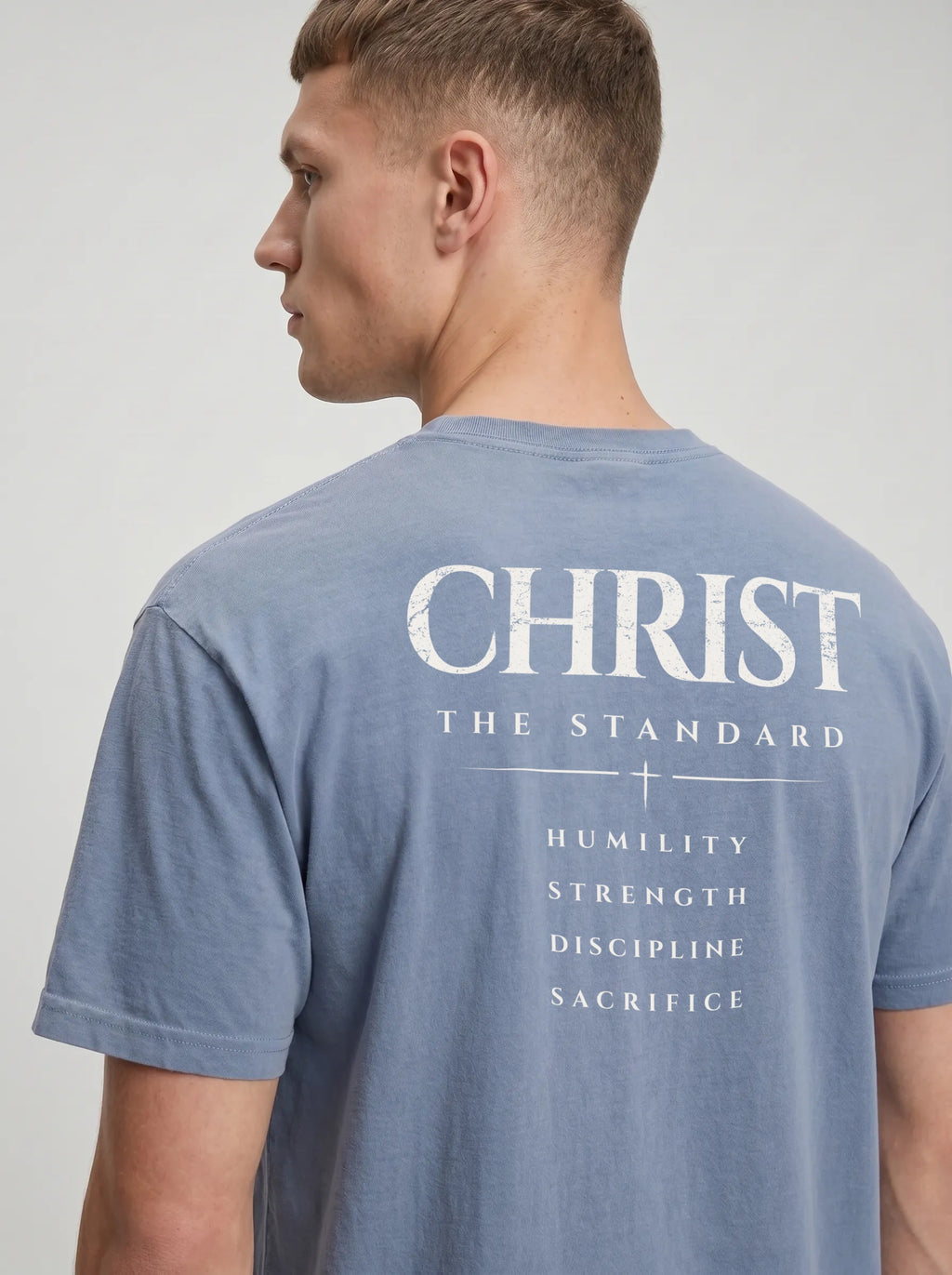 Angled back view of a person in a faded blue T-shirt reading “Christ The Standard,” with a cross and “Humility, Strength, Discipline, Sacrifice.”