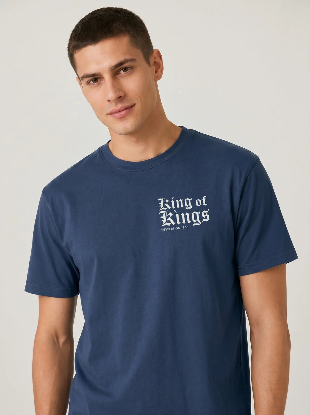 A man wearing a navy blue crewneck t-shirt with 'King of Kings' printed in white Gothic lettering on the chest, photographed against a neutral light gray background.