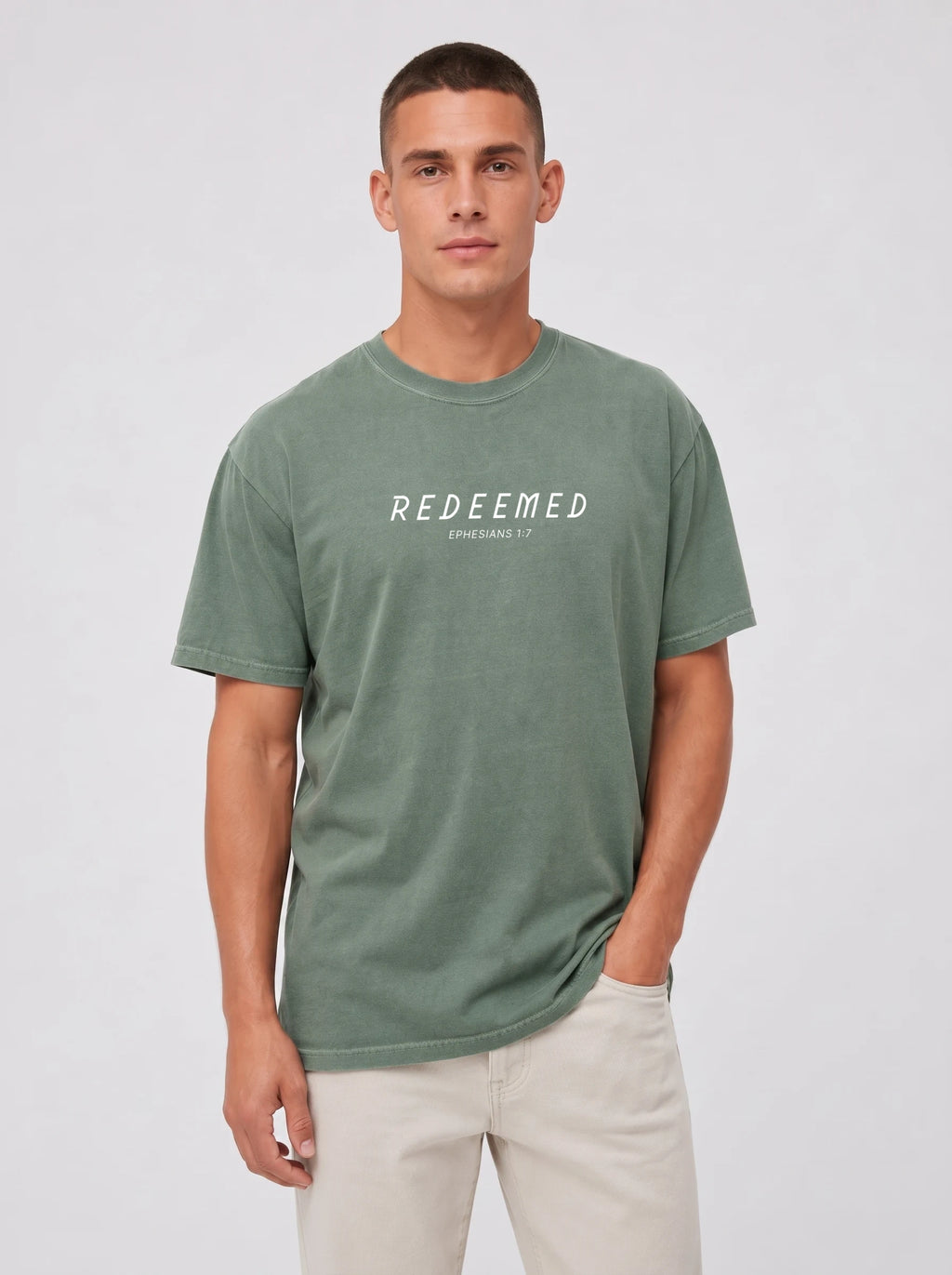 A man wearing a sage green Comfort Colors t-shirt with "REDEEMED" in bold white capital letters and "EPHESIANS 1:7" underneath, paired with beige pants, against a plain light gray studio background.