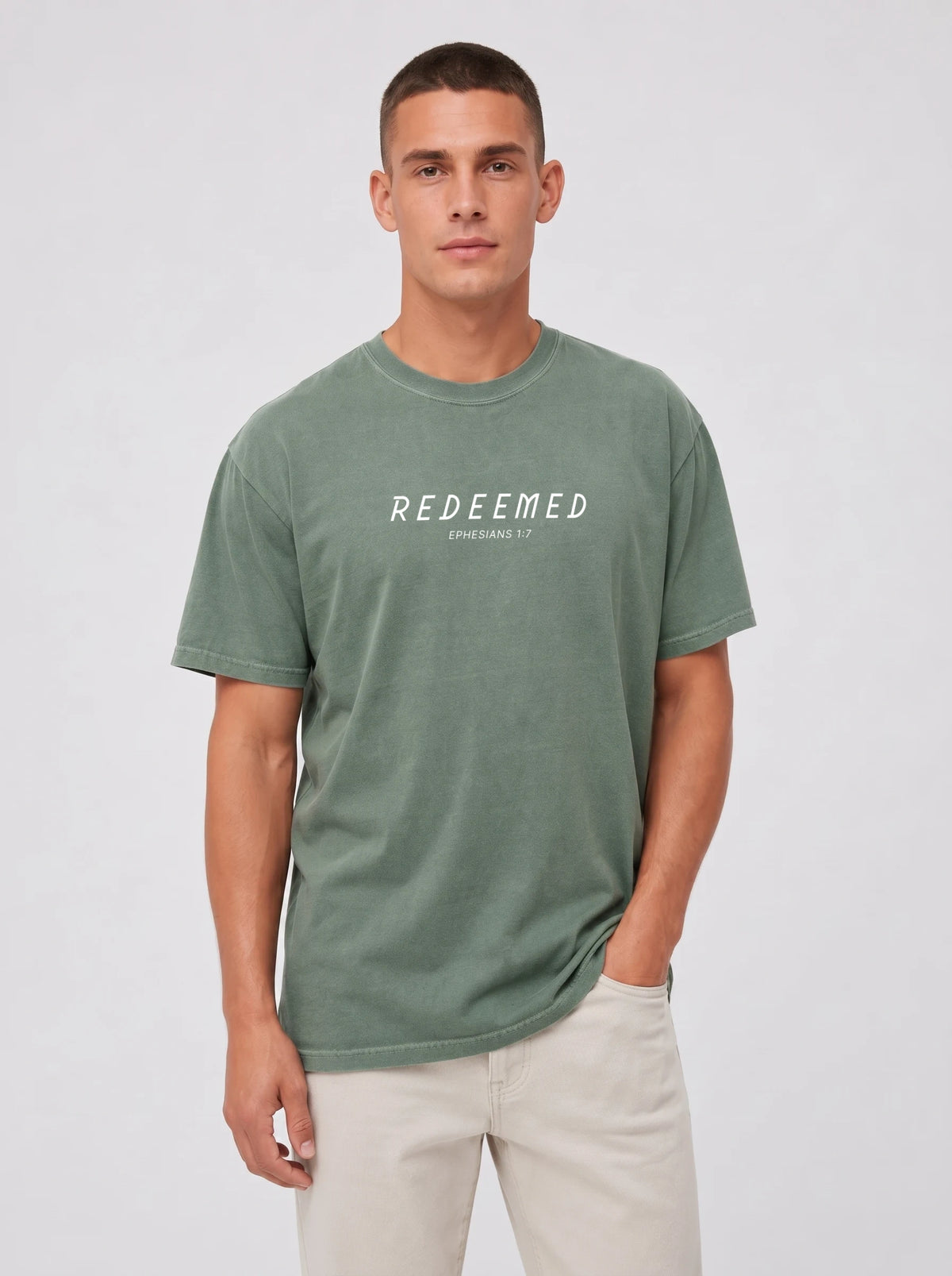 A man wearing a sage green Comfort Colors t-shirt with "REDEEMED" in bold white capital letters and "EPHESIANS 1:7" underneath, paired with beige pants, against a plain light gray studio background.