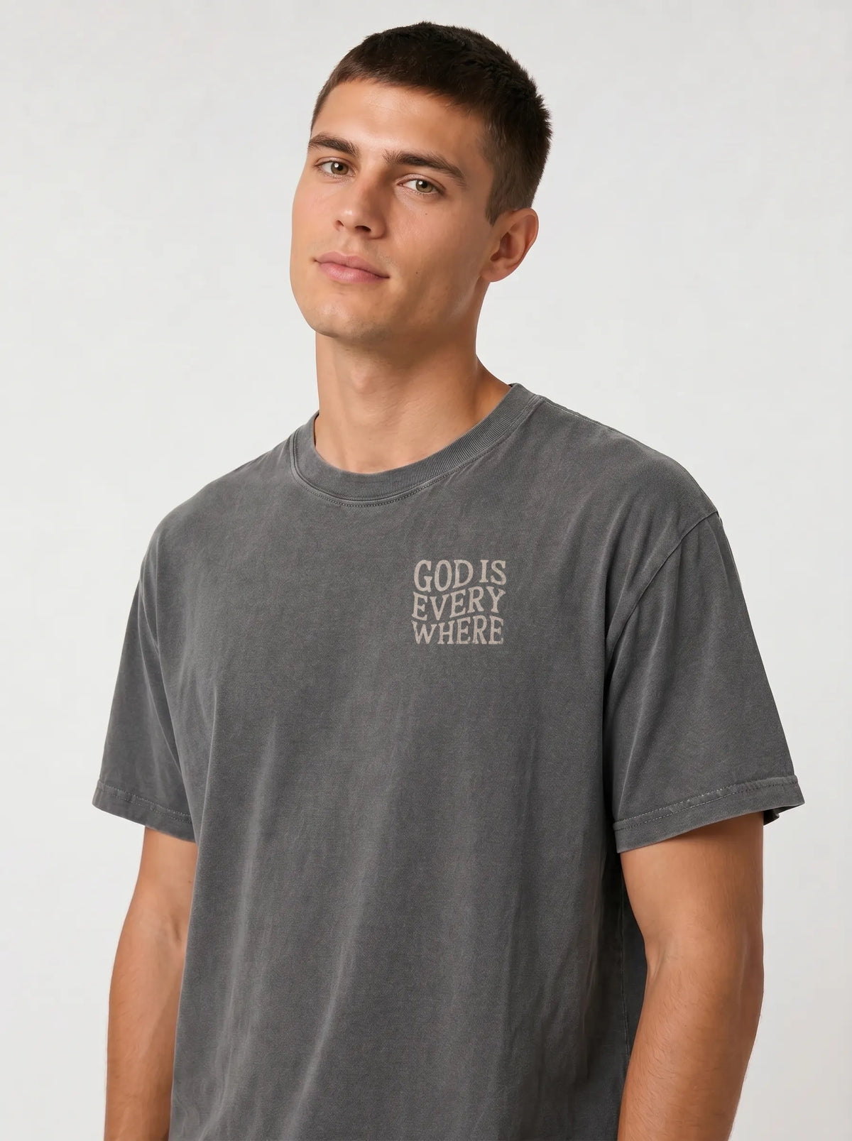 A man wearing a dark gray washed t-shirt with the text 'GOD IS EVERY WHERE' printed in white on the left chest, paired with light blue jeans, against a light gray studio background.