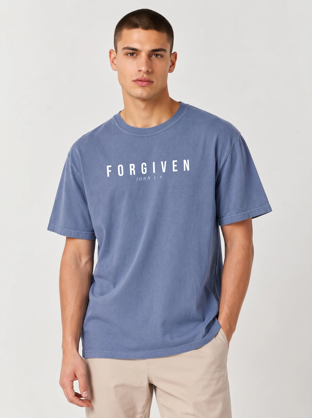 A man wearing a slate blue t-shirt with the word 'FORGIVEN' and 'JOHN 1:9' printed in white across the chest, paired with beige pants, against a light gray studio background.