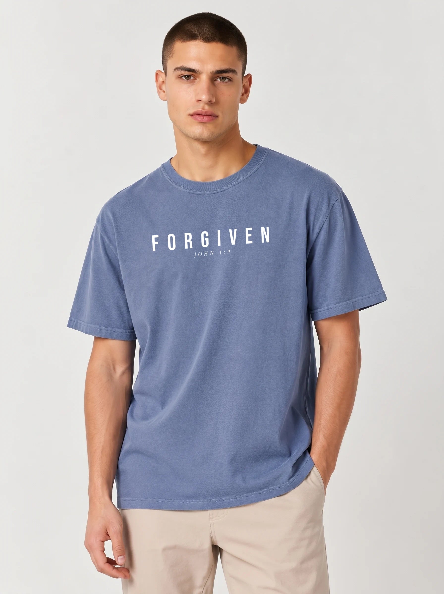 A man wearing a slate blue t-shirt with the word 'FORGIVEN' and 'JOHN 1:9' printed in white across the chest, paired with beige pants, against a light gray studio background.